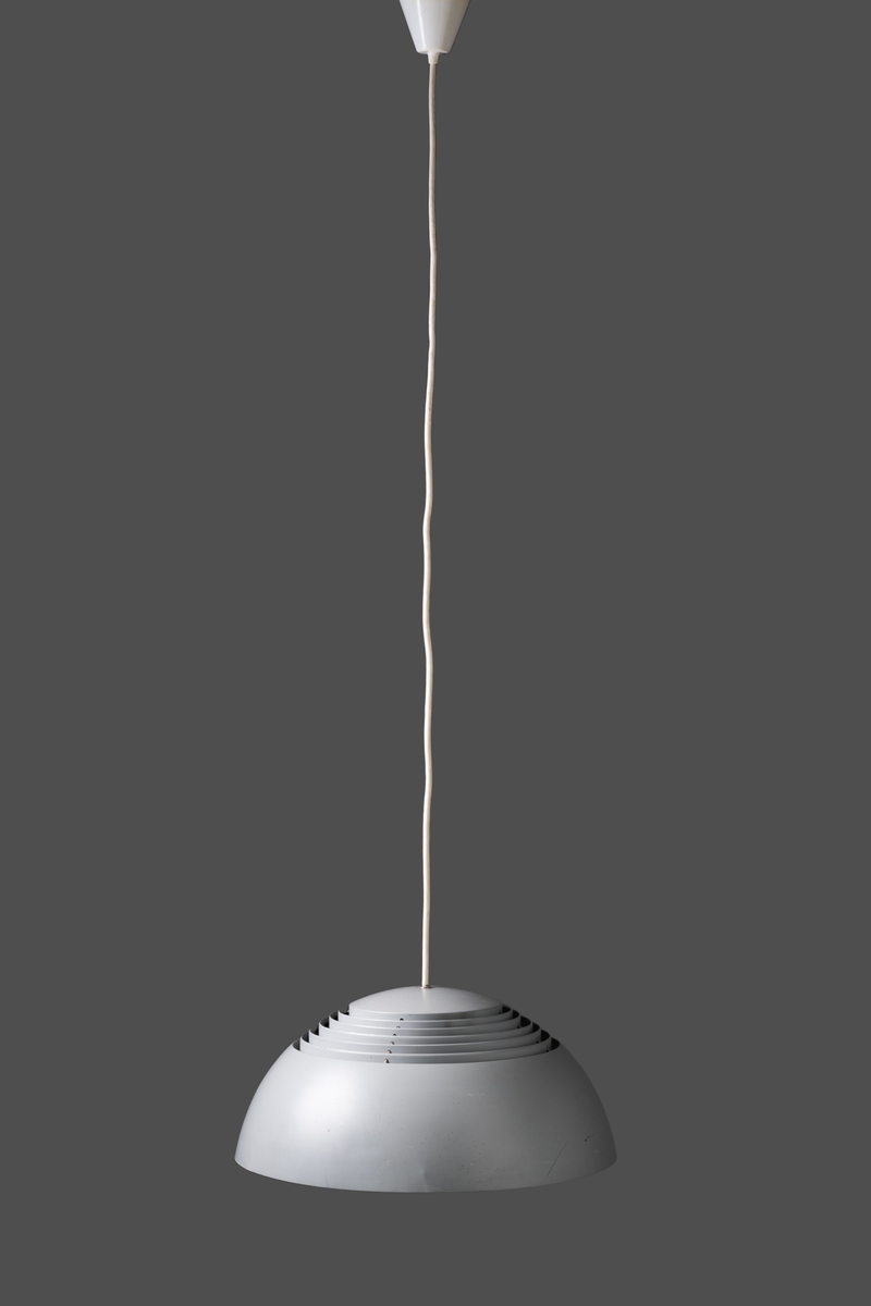 Taklampa av lackerad plåt. Lampan har en yttre halvklotformad kupolskärm med fem lameller på toppen för svagt uppåtriktat ljus. Den har invändigt tre glödlampssocklar. Skärmen är grå utvändigt och vit invändigt. Den inre, mindre skärmen är helt täckt och helt vitlackerad samt försedd med en glödlampssockel. Vit plastsladd utan kontakt, och vit takkopp av plast.