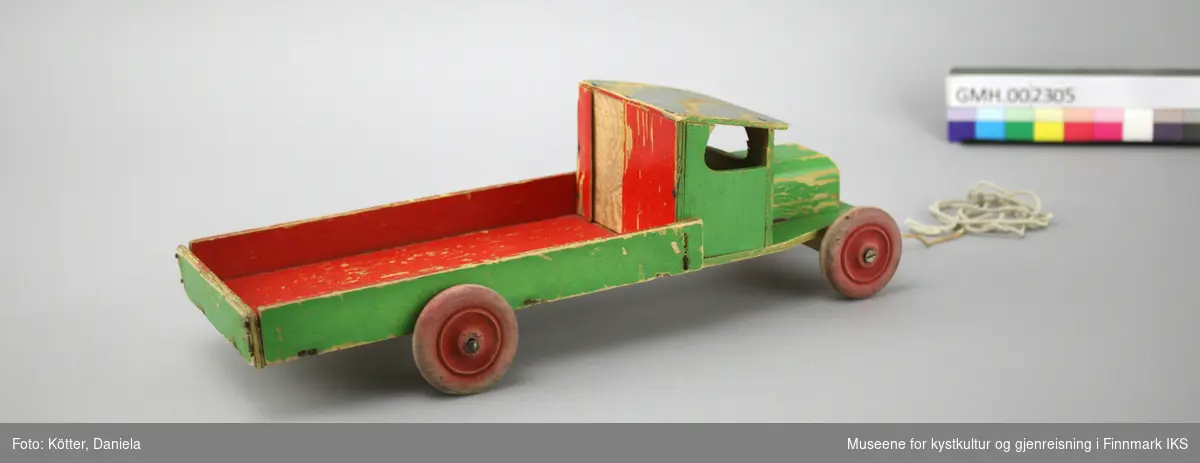 Lekebilen er laget av tre og kryssfinér. Den er malt grønn og rød. Bilen har en trekksnor festet i panseret