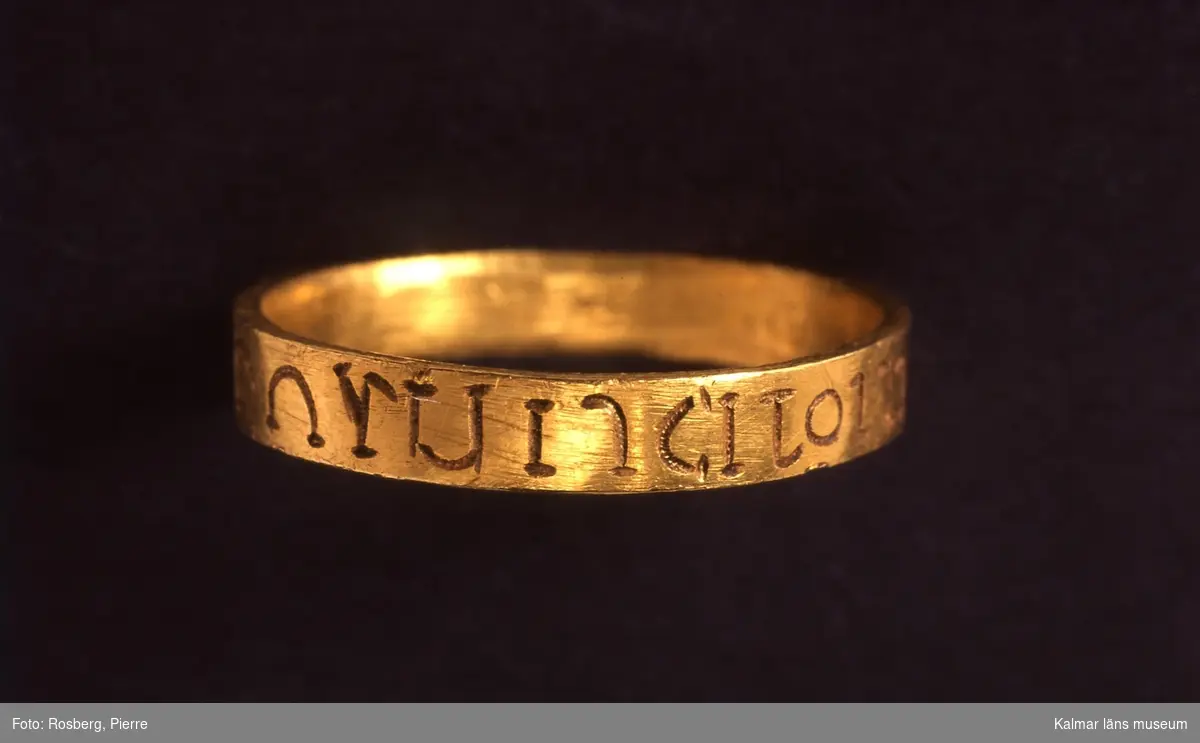 KLM 15873. Fingerring, bandformig av guld. Grekisk inskription runt hela ringen. (AMOR OMNIA VINCIT enligt B. Almgren 1948) Ska vara:  Amor vincit omnia. Förlovningsring? Datering: 1100-tal.