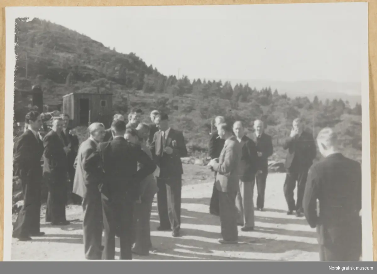 Utendørsmotiv. En gruppe menn i dress står på en grusvei eller -slette. Spredt skogvekst og en hytte eller uthus i bakgrunnen. Fotografert i forbindelse med Vestlandsk Trykkerstevne i Bergen, 1946.