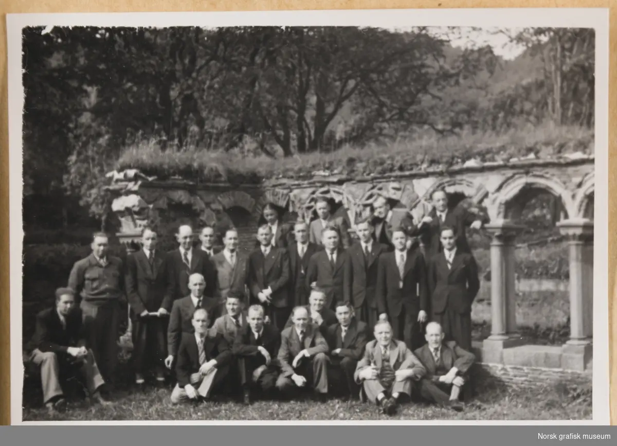 Utendørsmotiv. Tjuefem menn i dress og slips oppstilt foran en søylegang i stein med gress på toppen. Fra Lysekloster ved Bergen. Fotografert i forbindelse med Vestlandsk Trykkerstevne i Bergen, 1946.