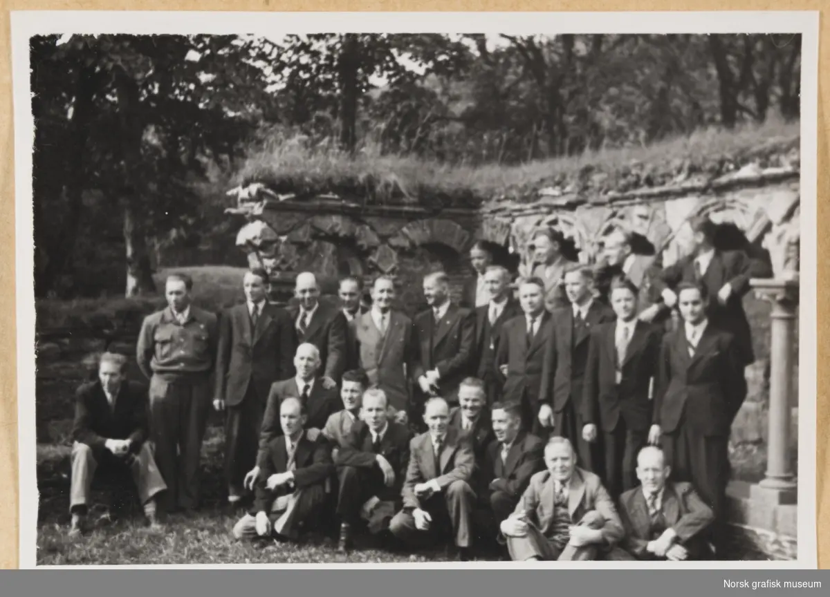 Utendørsmotiv. Tjuefem menn i dress og slips oppstilt foran en søylegang med gress på toppen. Fra Lysekloster. Fotografert i forbindelse med Vestlandsk Trykkerstevne i Bergen, 1946.