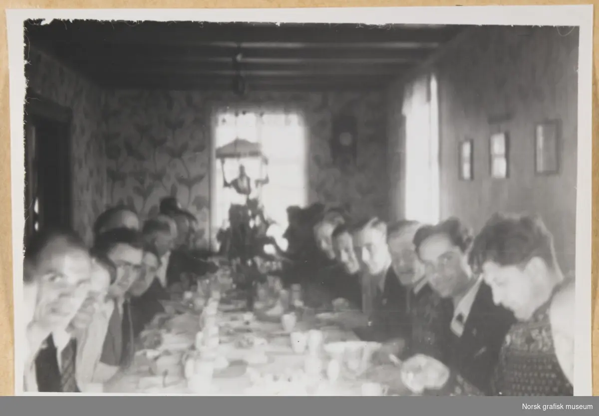 Innendørs, i en stue med storblomstret tapet på veggene og lysekrone i taket. Mange mennesker sitter ved et langbord dekket med diverse servise. Fotografert i forbindelse med Vestlandsk Trykkerstevne i Bergen, 1946.