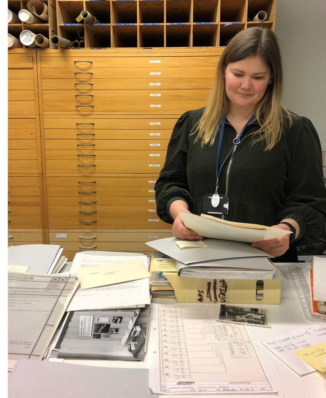 Bildet viser en ung kvinne som står ved et bord fylt av papirer. Hun ser ned på noe dokumenter hun holder i hendene.