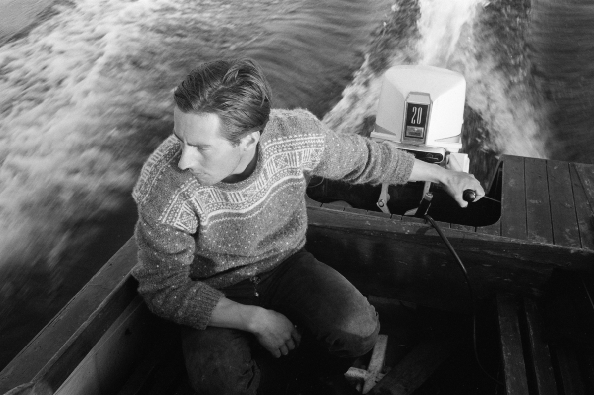 Mann i strikkegenser styrer båt med påhengsmotor.
