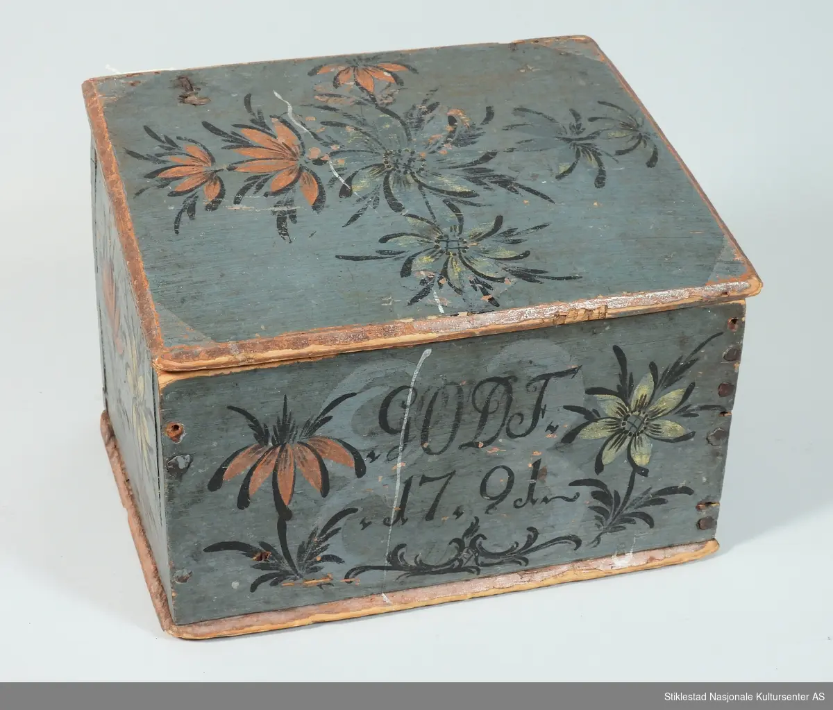 Blåmalt lite skrin med skrått lokk. Rosemalt på tre sider, samt lokk. Teksten og konturen av blomster er svartmalt. Blomster i rød og gulhvit farge. Rødmalte kanter på skrin. Lokket er malt hvitt på innsiden, der er det malt en stilisert blomst i orange farge. Naglet sammen med jernspiker. Hengslet lokk. Malt av Ole Haldosen Berg (1747-1828)