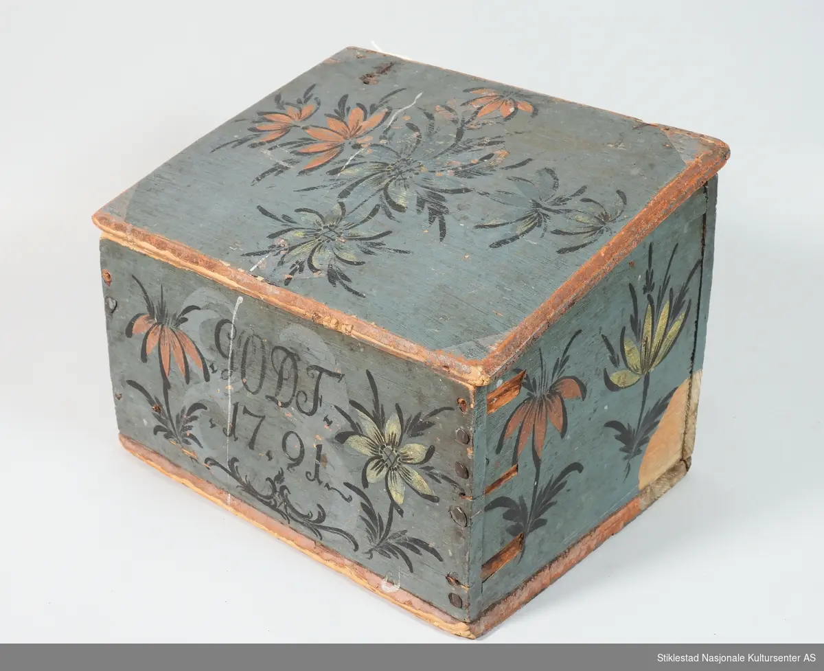 Blåmalt lite skrin med skrått lokk. Rosemalt på tre sider, samt lokk. Teksten og konturen av blomster er svartmalt. Blomster i rød og gulhvit farge. Rødmalte kanter på skrin. Lokket er malt hvitt på innsiden, der er det malt en stilisert blomst i orange farge. Naglet sammen med jernspiker. Hengslet lokk. Malt av Ole Haldosen Berg (1747-1828)