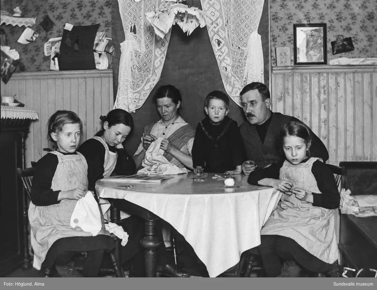 Alma och Gottfrid Höglund med barnen Hilma, Ingrid, Irma och Per. Familjen sitter runt ett bord och är sysselsatta med olika göromål.