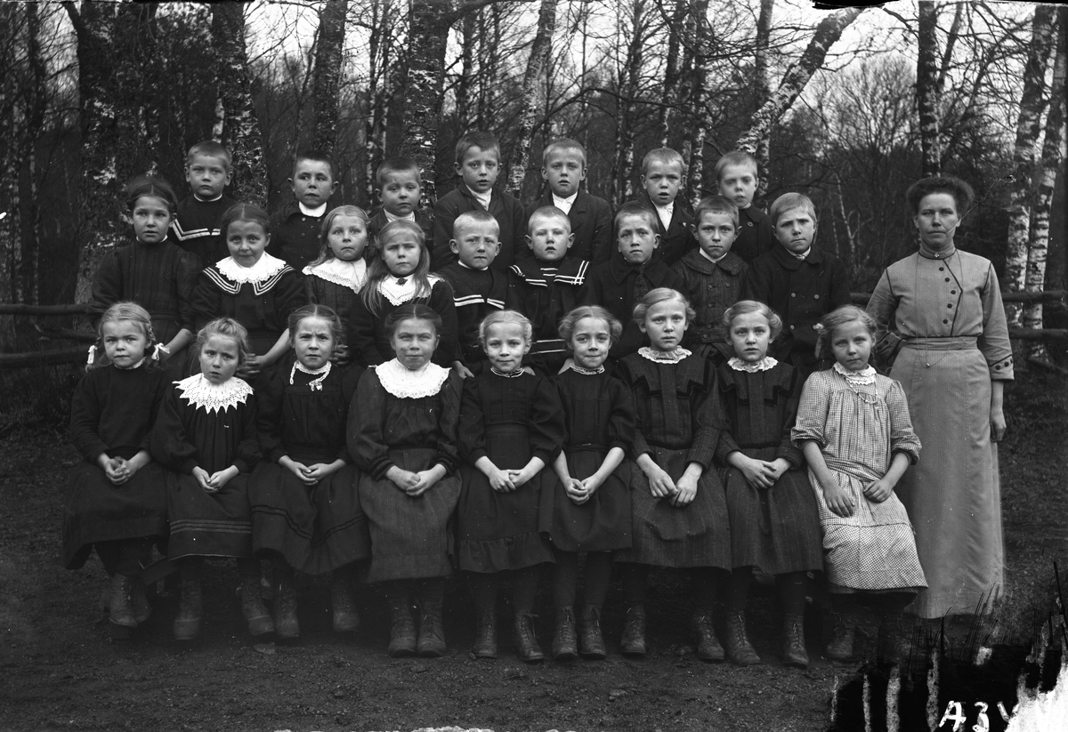 En skolklass med 25 barn och lärarinnan till höger. Barnen är mörkt klädda, utom en flicka som har en ljus, rutig klänning. De är uppställda utomhus framför en björkdunge.