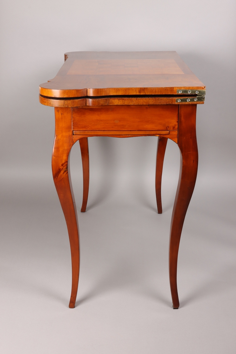 The card table has a double top: one for chess and one for card playing. The frame conceals a concertina mechanism which extends the frame to support the folded table. Unfolded the frames inside has a textile backgammon board. The double top is veneer in dark wood, possibly burr walnut and the carcase wood is pine or alder tree.