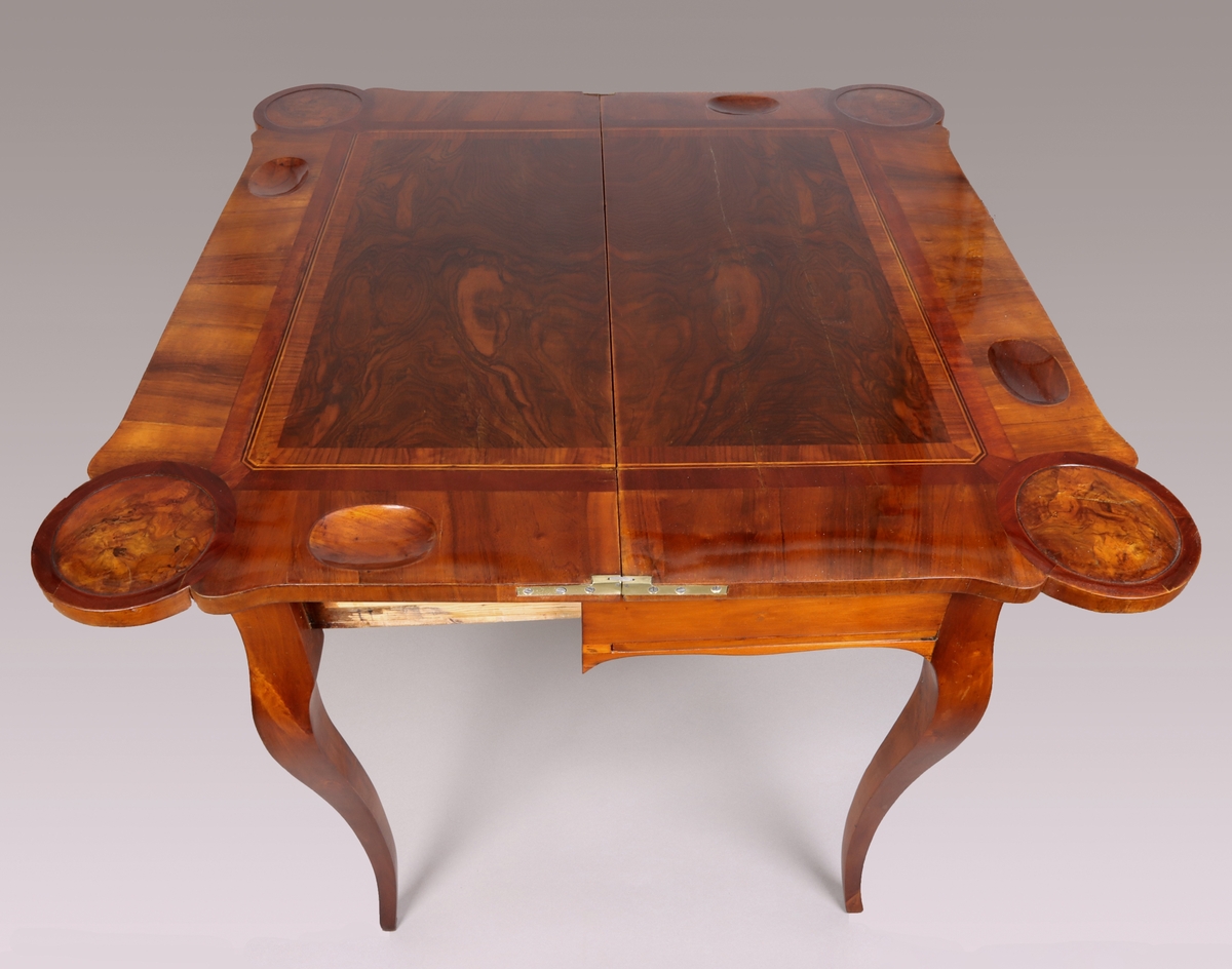The card table has a double top: one for chess and one for card playing. The frame conceals a concertina mechanism which extends the frame to support the folded table. Unfolded the frames inside has a textile backgammon board. The double top is veneer in dark wood, possibly burr walnut and the carcase wood is pine or alder tree.