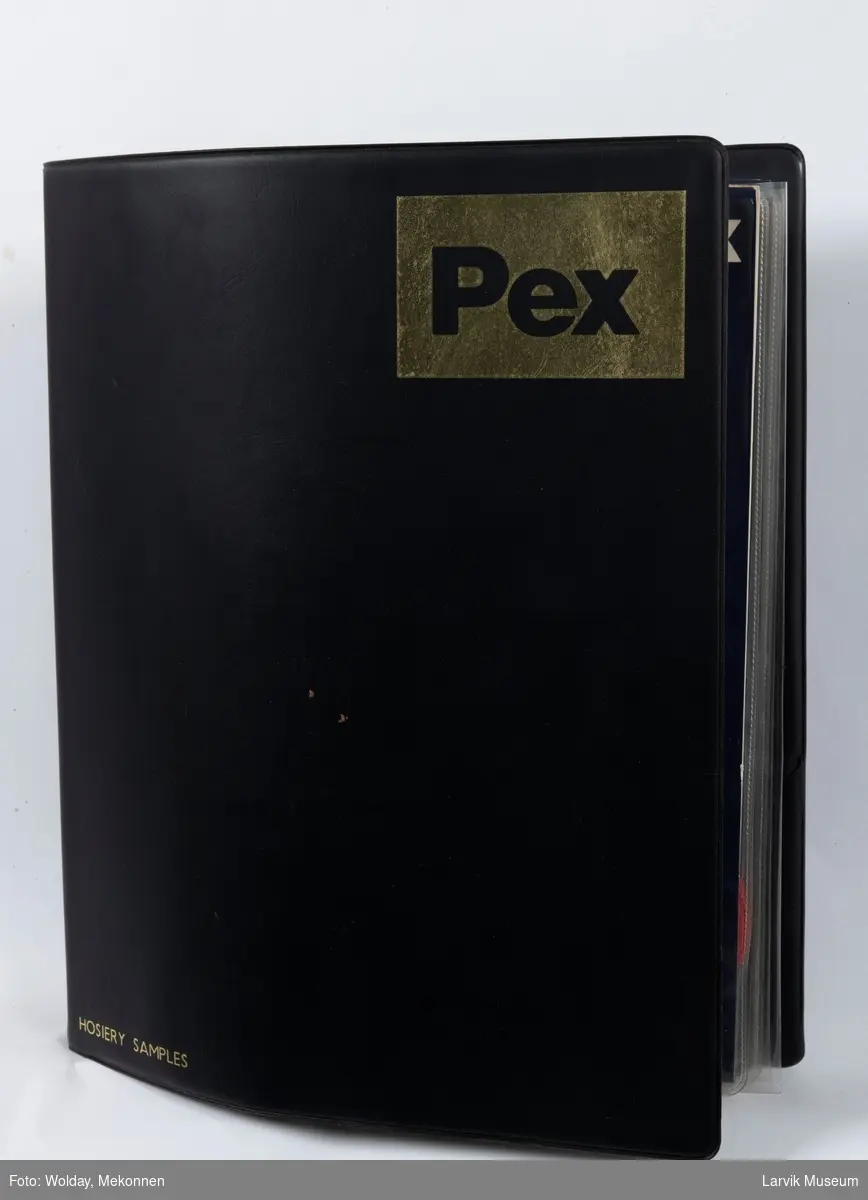 På forsiden av permen står det: Hosiery samplex  merke:PEX 18 stk. barnesokker pluss noen stoffprøver ligger i en sort plastperm