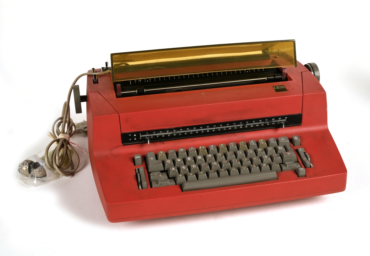 En elektrisk skrivemaskin fra IBM med kulehode og rettetast. Kassen til skrivemaskinen er av rød plast. Plasten er litt ruglete i overflaten. Tastene er av grå plast med hvit påskrift. Over rommet man setter i ark er det et lokk av grålig, gjennomsiktig plass.