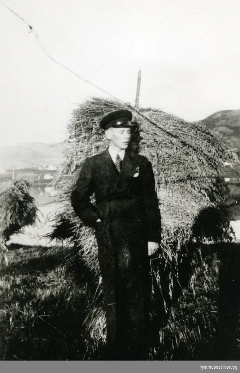 Ung ukjent mann i dress og kasjettlue foran en kornstaur i Flatanger