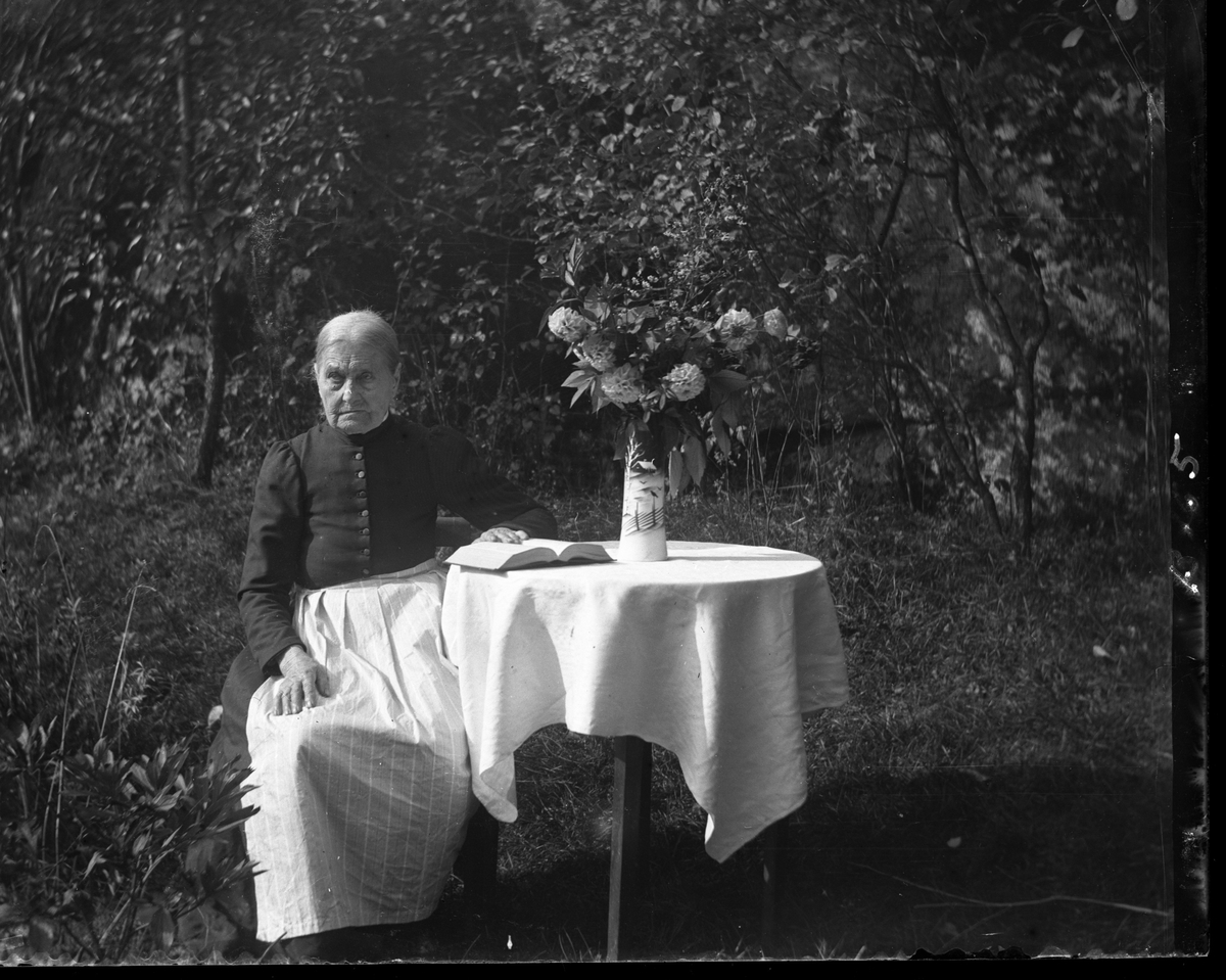 En äldre dam med förkläde sitter utomhus vid ett dukklätt bord. Hon håller handen på en bok på bordet och bredvid står en bukett dahlior i en hög vas.