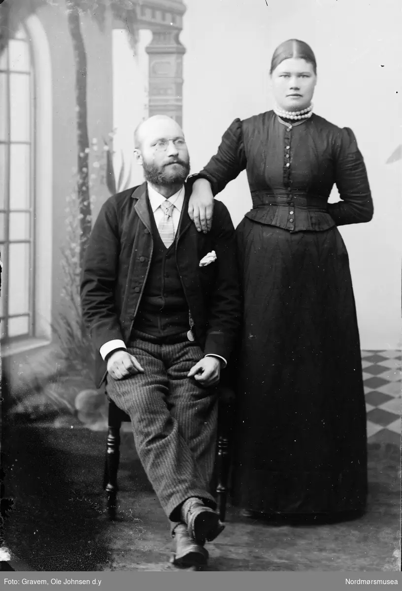 Portrett av kvinne og mann i helportrett. Kvinnen har på seg kjole og smykke og har hånden sin på skuldra til den sittende mannen.