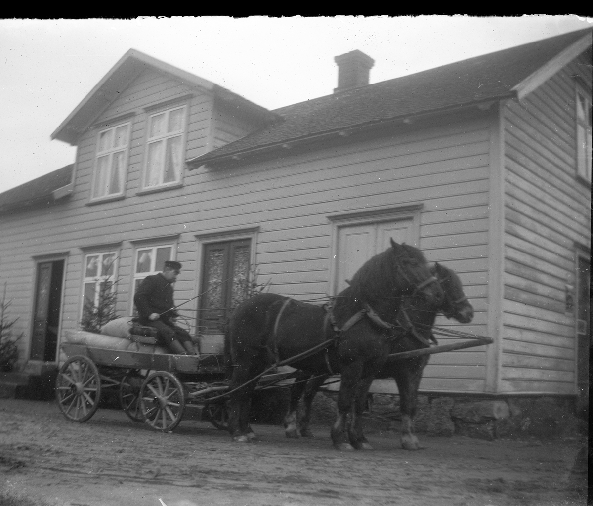 En man på kärra med säckar, dragen av två hästar. Ekipaget står utanför handelsboden i Sällstorp, Karlsberg. Kanske är det omkring jul eftersom det står flera granar utmed fasaden.