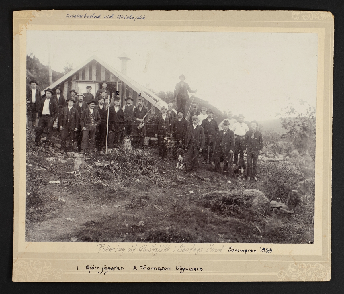 Rallarlag vid Abiskojokk i Söndagsskrud. Sommaren 1899. Grupporträtt framför arbetarbostad, två män är utmärkta: "Björnjägaren" och "Thomason Vägvisare".