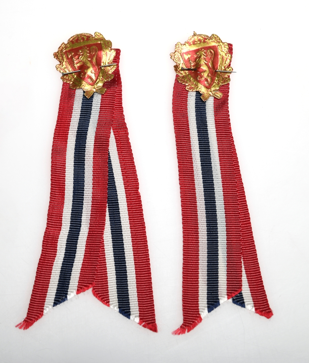 To identiske 17.mai-sløyfer. De er laget av silkebånd med striper i rødt, hvitt og blått. Båndene er brettet dobbelt og klippet skrått i endene. Oppe på sløyfene er det norske riksvåpenet festet med en knappenål. Riksvåpenet er laget av papir.