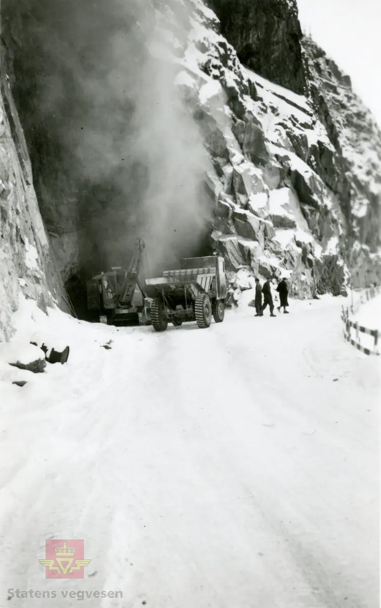 Tunnelarbeid i Kvamskleiva 1956-1957. 
En Ruston-Bucyrus wiregravemaskin laster masse over på en dumper med et litt spesielt førerhus ved tunnelinnslaget.