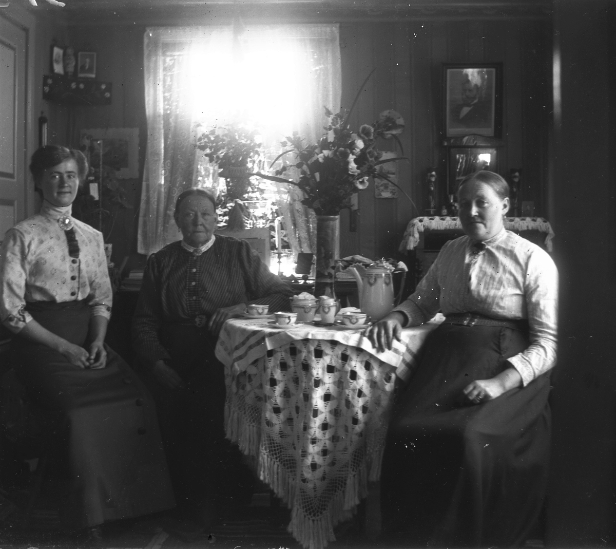 Tre kvinnor dricker kaffe vid ett runt bord. De kan vara ur tre generationer i huset och är alla klädda i blus med brosch halsen, långskjolar och skärp. En stor blombukett i hög vas pryder bordet. De har en rullgardin nerdragen med ett landskapsmotiv.
(Se även bildnr GEB199)