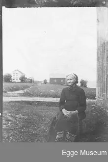 Kristianne Nilsdtr. Mortensen (1823-1918) sitter på en stol/krakk utenfor en bygning. 