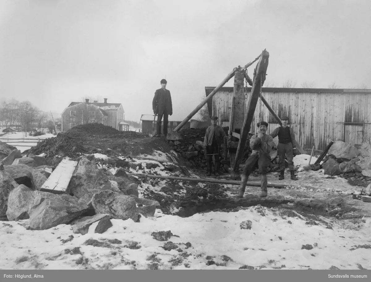 Fyra arbetsklädda män vid någon slags kran/lyftanordning, möjligen för att lyfta sten. I bakgrunden en mangårdsbyggnad och andra byggnader.