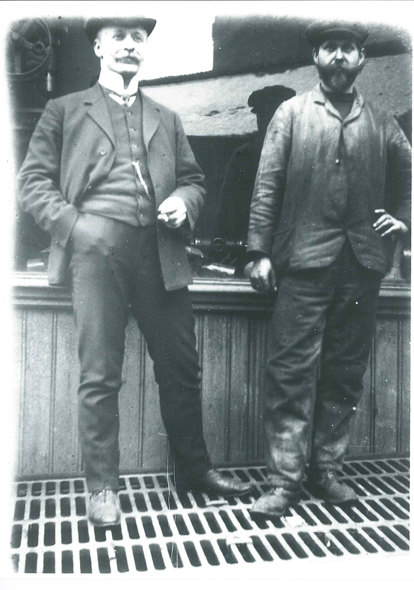 Mann i "borgerlig" klesdrakt, skjorte med høysnipp, vest med urkjede, og skalk, står ved siden av mann i arbeidsklær.