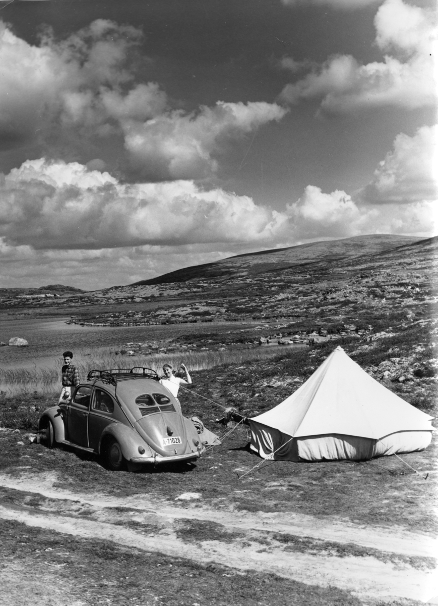 Foto av et ungt par som har satt opp et telt, delvis festet i en bil av merket Volkswagen "Boble". 