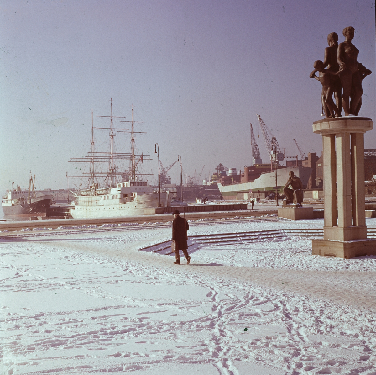 En mann går på Rådhusplassen vinteren 1962-63. I bakgrunnen ses Akers mekaniske verksted og Båter som ligger ved Rådhuskaia. En av Emil Lies skulpturer sees til høyre i bildet.