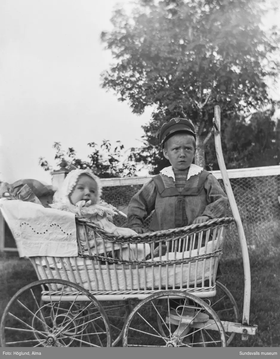 Två små barn utomhus, den ena sitter i en rottingvagn med stora hjul och draghandtag.