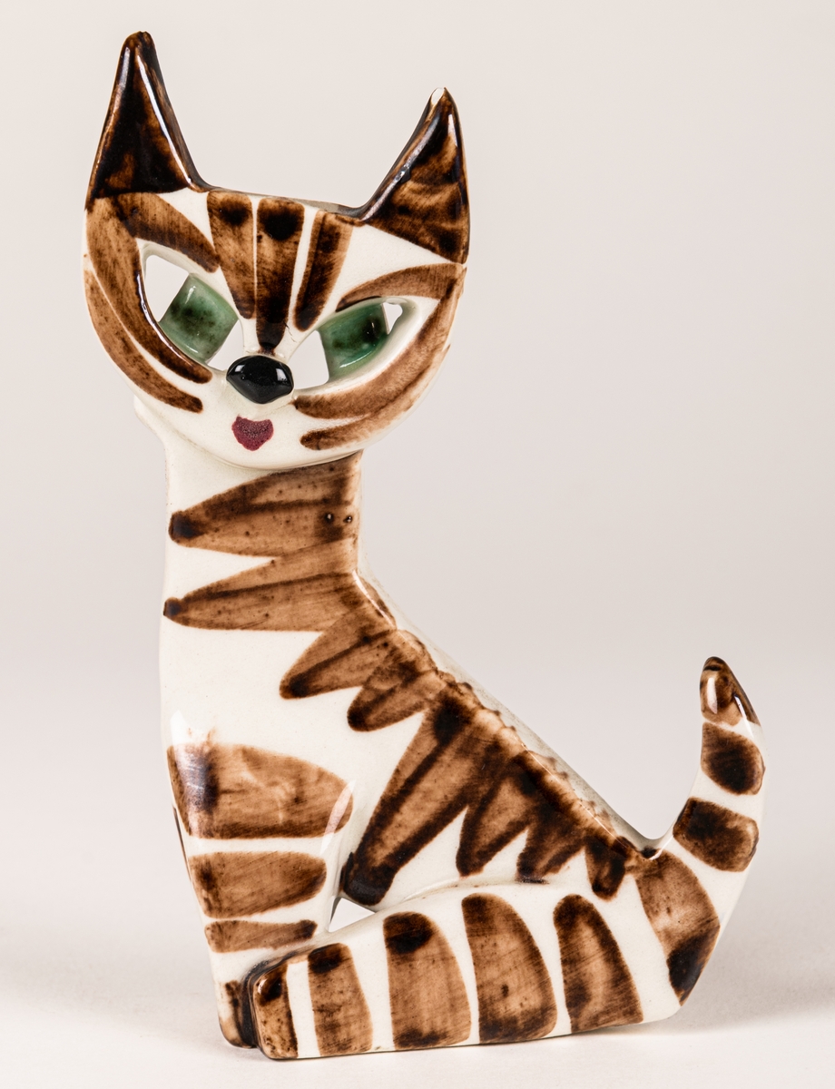 Figurin i form av en katt, kavlad och skuren flintlera, glaserad med brun dekor föreställande päls, gröna ögon. Formgiven av Dorothy Clough och tillverkade 1955-1962.