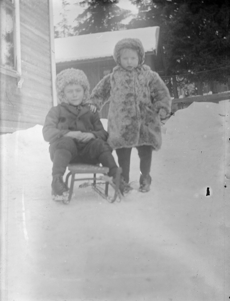 Portrett av to barn. Bildet er tatt utendørs om vinteren, det største barnet sitter på en kjelke.