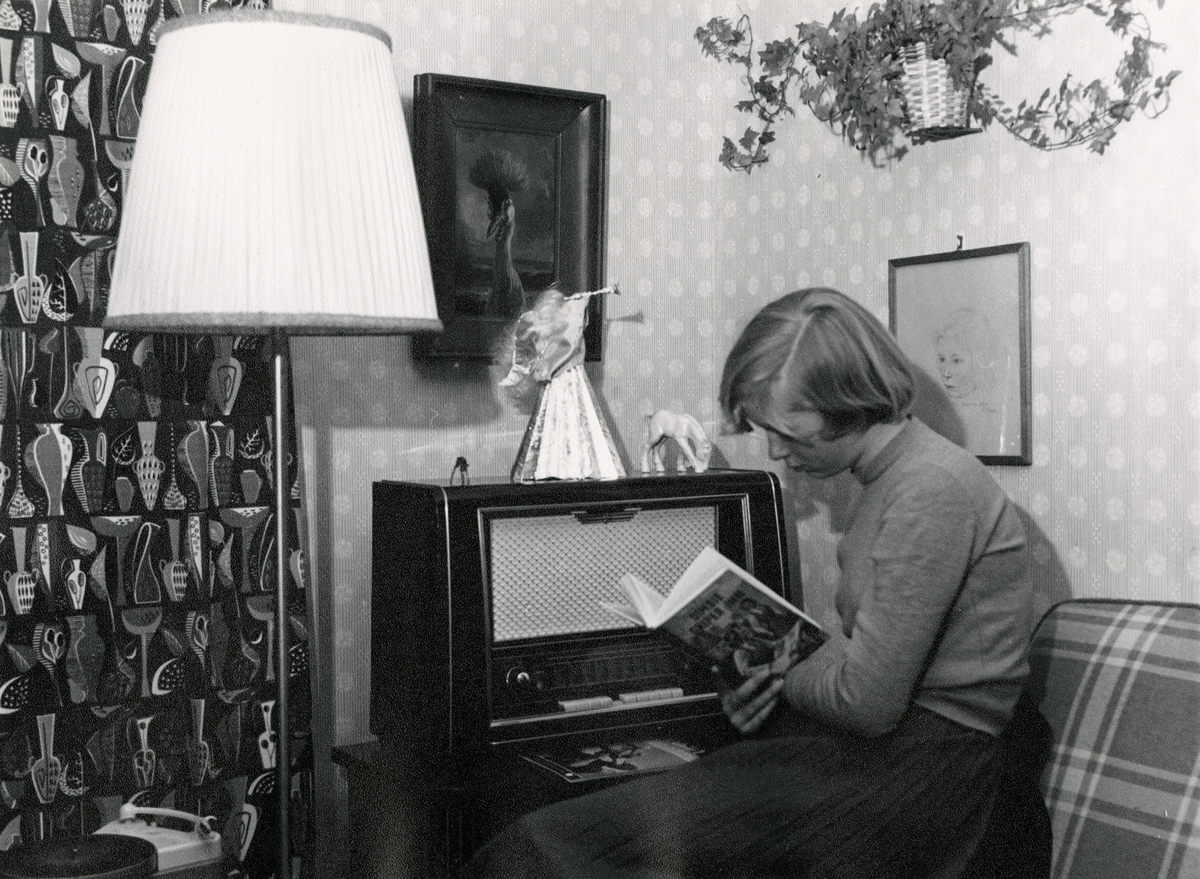 En ung jente sitter og leser "Dimsie griper inn" ved siden av en radio/ kabinett.  