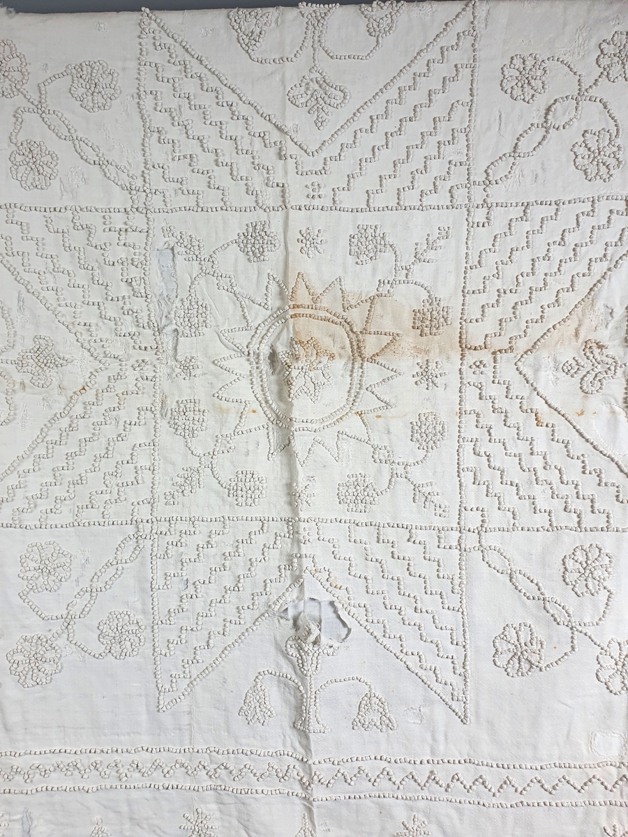 Hvitt, fôret sengeteppe av bomull, med brodert mønster. Teppet er lappet og har noen hull, og har noen flekker.