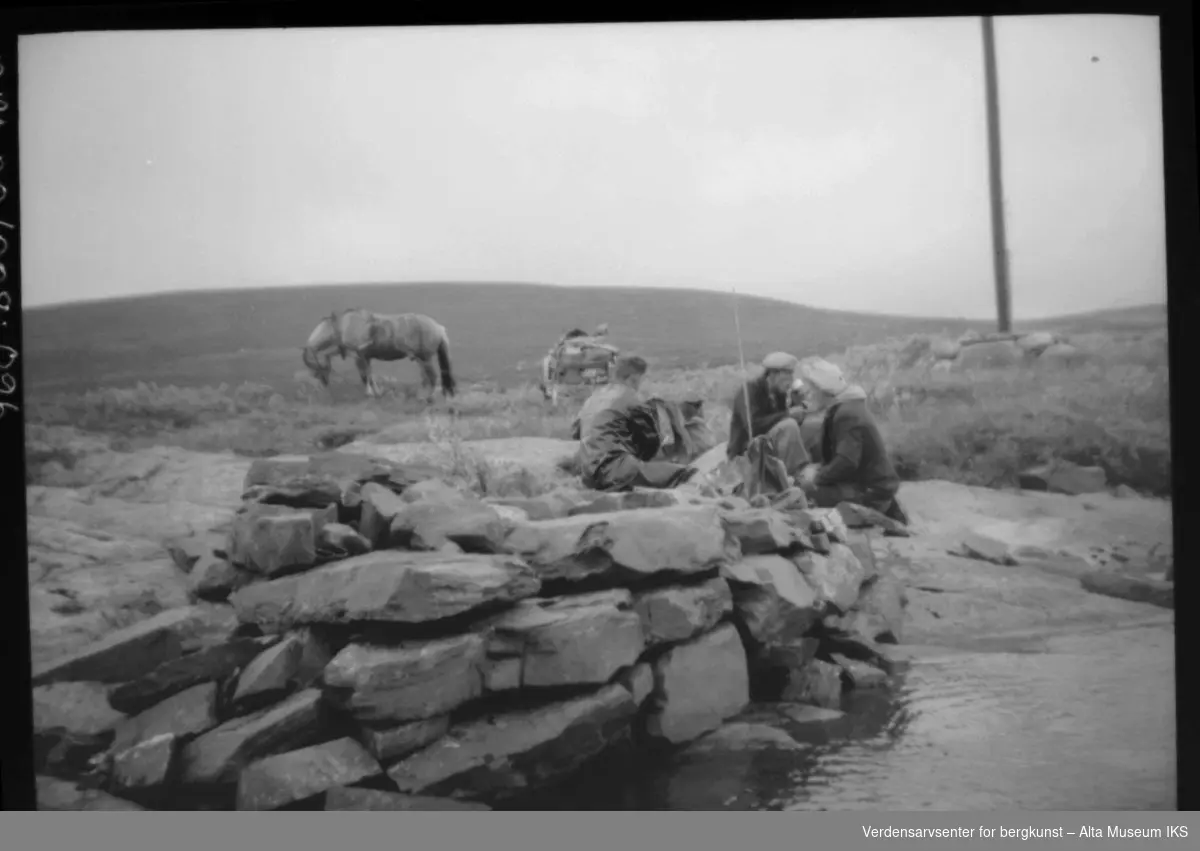 3 menn med hest, fiskestang og ryggsekk sitter ved en steinmur i vannkanten.