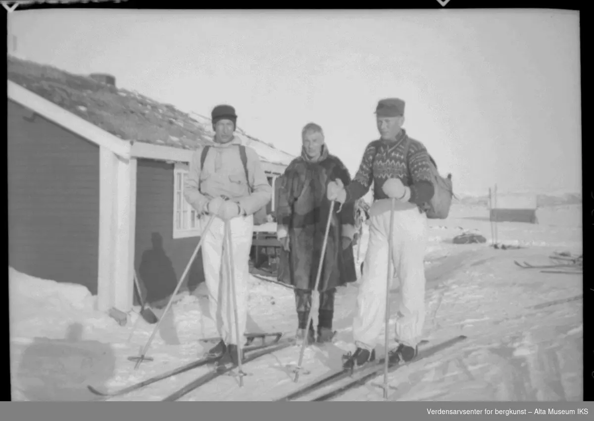 Tre menn foran ei hytte ved Joatka fjellstue. To menn har på skiutstyr, den tredje er kledd i pesk, bellinger og skaller.