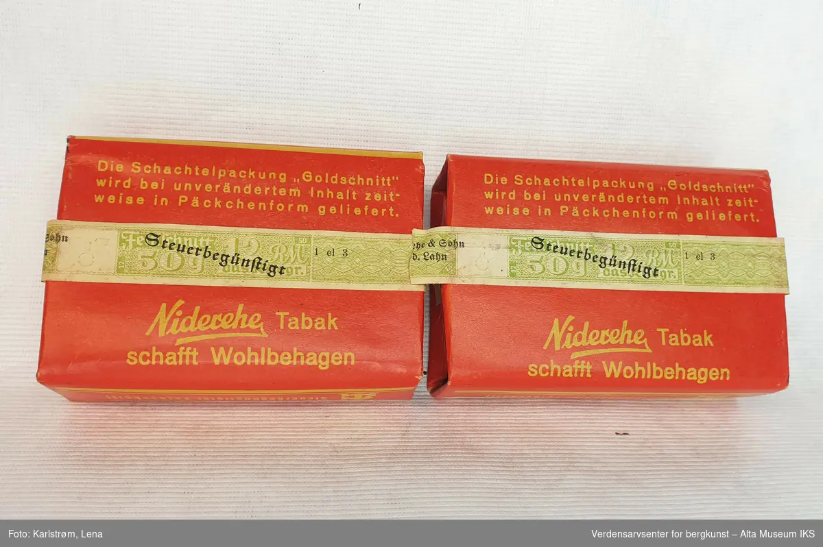 To tobakksesker med tobakk innpakket i rødt papir. Eskene har gul tekst og illustrasjon av tobakksblader. Eskene er forseglet med magebelte, stemplet med hakekors og den tyske ørn.