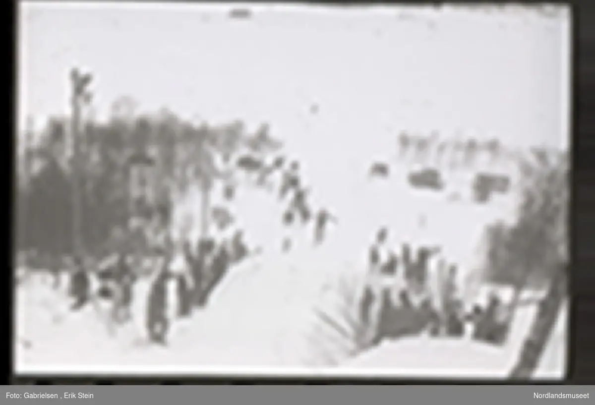 Fotografi av et skihopp som det hopper et barn med ski på seg fra mens noen kvinner og menn og barn med ski og skistaver og vinterklær og deltakernummer på seg som står og ser på barnet som hopper fra skihoppet vedsiden av skihoppet og lenger nede i bakken og tre barn som står i et utsiktstårn 
og ser på skihopperen og noen biler og busser som står parkert lenger nede i skibakken vedsiden av en vei som det vokser noen trær vedsiden av og man ser en skiløper ute i snømarken som har nettopp landet i snøen etter å ha hoppet fra skihoppet