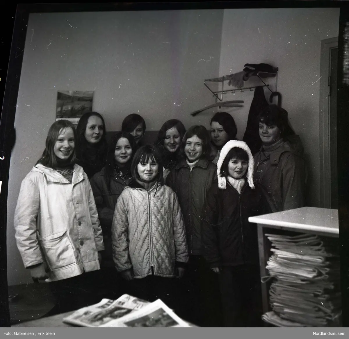 Fotografi av ti jenter kledd i vinterklær som står i et rom 
med et bord og en hylle som det ligger mange aviser på 
og en kalender som henger på veggen 
og en hylle med et kleshengestativ til og henge klærene sine på som det henger en genser på og ligger noen votter 
og et skjerf på