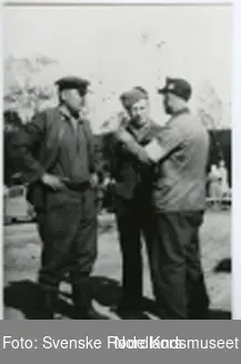 Fotografi av en krigsfange kledd i en gråsvart skjorte og bukse 
og støvler og en hatt på hodet og en krigsfange kledd i en gråsvart skjorte og bukse og en hatt på hodet som snakker med en lege kledd i en grå skjorte og svart bukse
og en hatt med et merke på hodet og et hvitt bånd med røde kors symbolet rundt armen til uniformen til legen som står i Klungset 
leir i nærheten av noen sykepleiere og biler i leiren
og snakker med de to krigsfangene