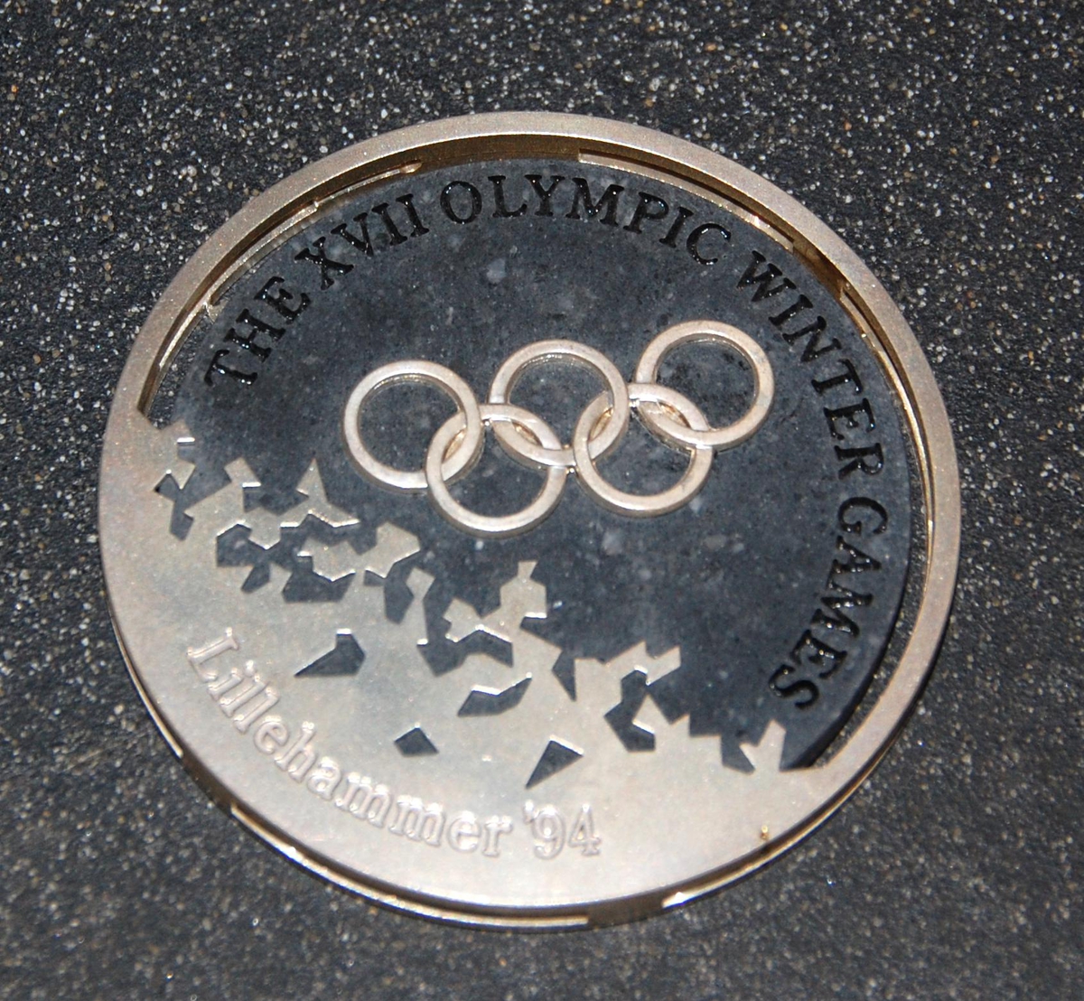 Medalje av metall og stein. På den ene siden er det motiv av de olympiske ringene over krystallmønster. På den andre siden er det piktogram av en skiløper og emblemet for de olympiske vinterleker på Lillehammer i 1994. Medaljen er plassert på et stativ av granitt.