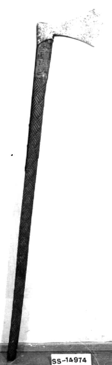 Asymmetrisk økseblad, sammensveiset med overliggende jernskinne.
Fremre øksebladspiss brukket.
Rikt dekorert skaft med flettet rutemotiv. Innskripsjon på øvre del av skaftet (se Påført tekst)
