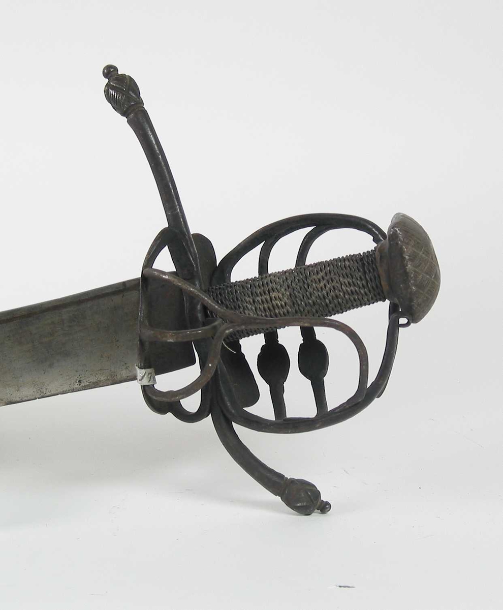 Pyramideformet knapp med strekdekor. Asymmetrisk kurvfeste med to sidebøyler( øverste avbrukket )med riflede fortykkelser på midten. mellom håndbøylen og kurvens ytterkant. Innsiden har en  sidebøyle. Kurvens basis er på utsiden en kvadratisk parérplate med to hjerteformede gjennombrytninger. På innsiden er det en klingebrekker og tommelbøyle. Opp og nedbøyde parérstenger med riflede fortykkelser på endene.
Tregrep med nyere jerntrådvikling.
Enegget krum klinge med 3 hull ipå klingspissen og en smal hulslipning på ryggsiden.Tre smedmerker på venstre side. 