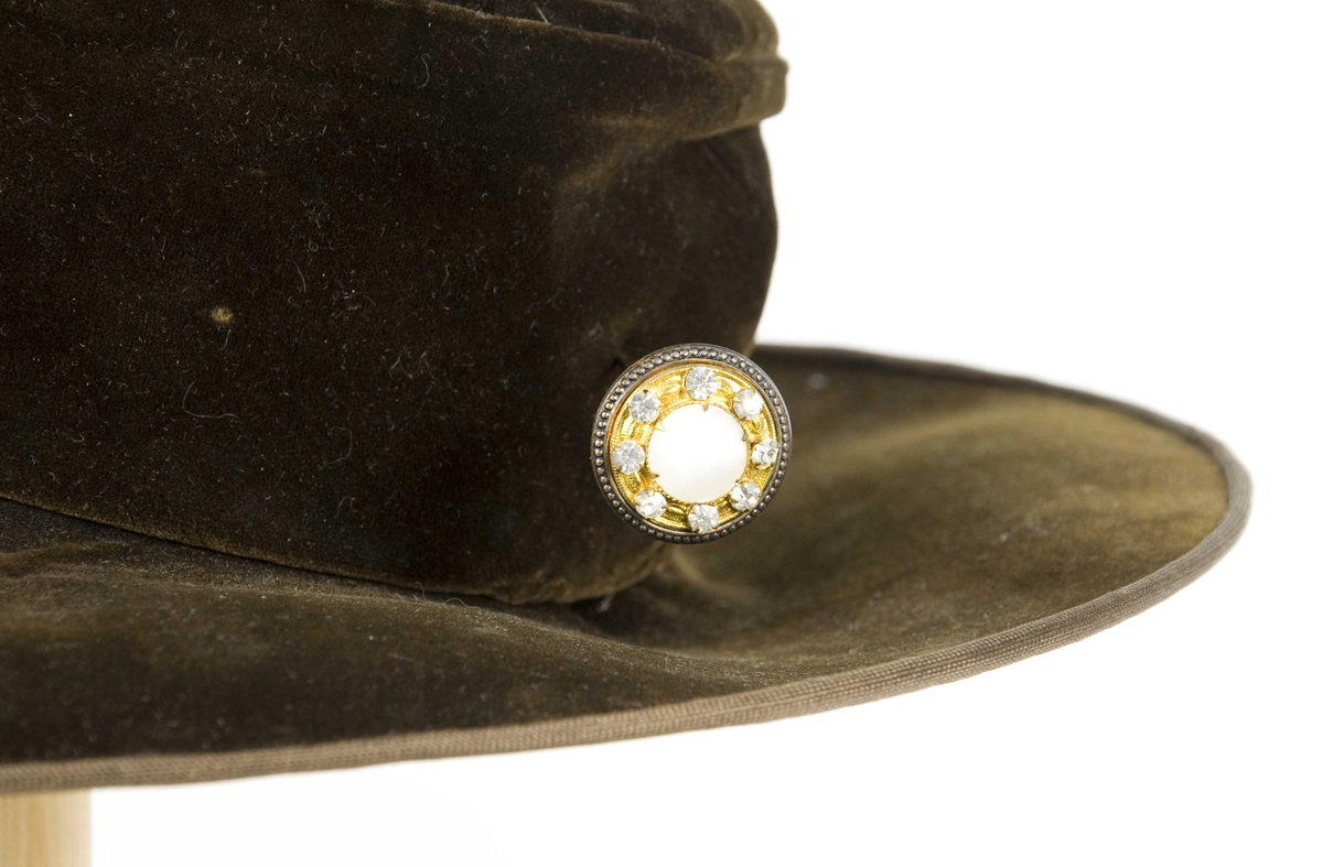 Grønn plysjhatt med høy pull og bred brem. hatten har et trefarget påsydd silkebånd som pynt på den ene siden (virker sekundær).  Midt bak en hattebnål. Fôr av silke.