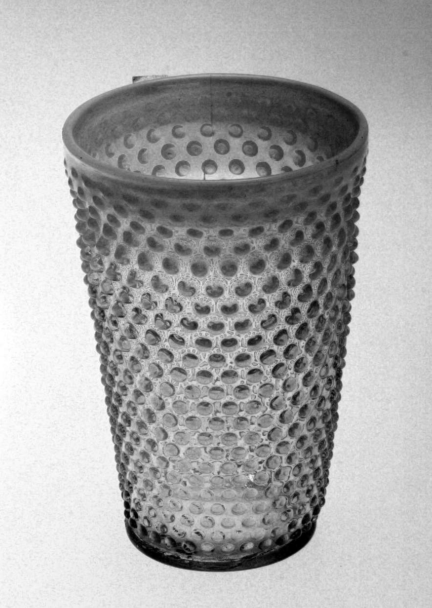 Blomstervase med konisk form. Den er i glass og yttersiden er 'knoppete'. Kanten av vasen og 'knoppene' har en melkehvit farge. Vasen er sprukket.