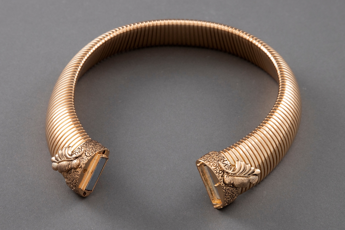 Halsklave i gullfarget metall. Smykket er bredt leddet med magnetlås midt foran med fjærdekor på låsen.