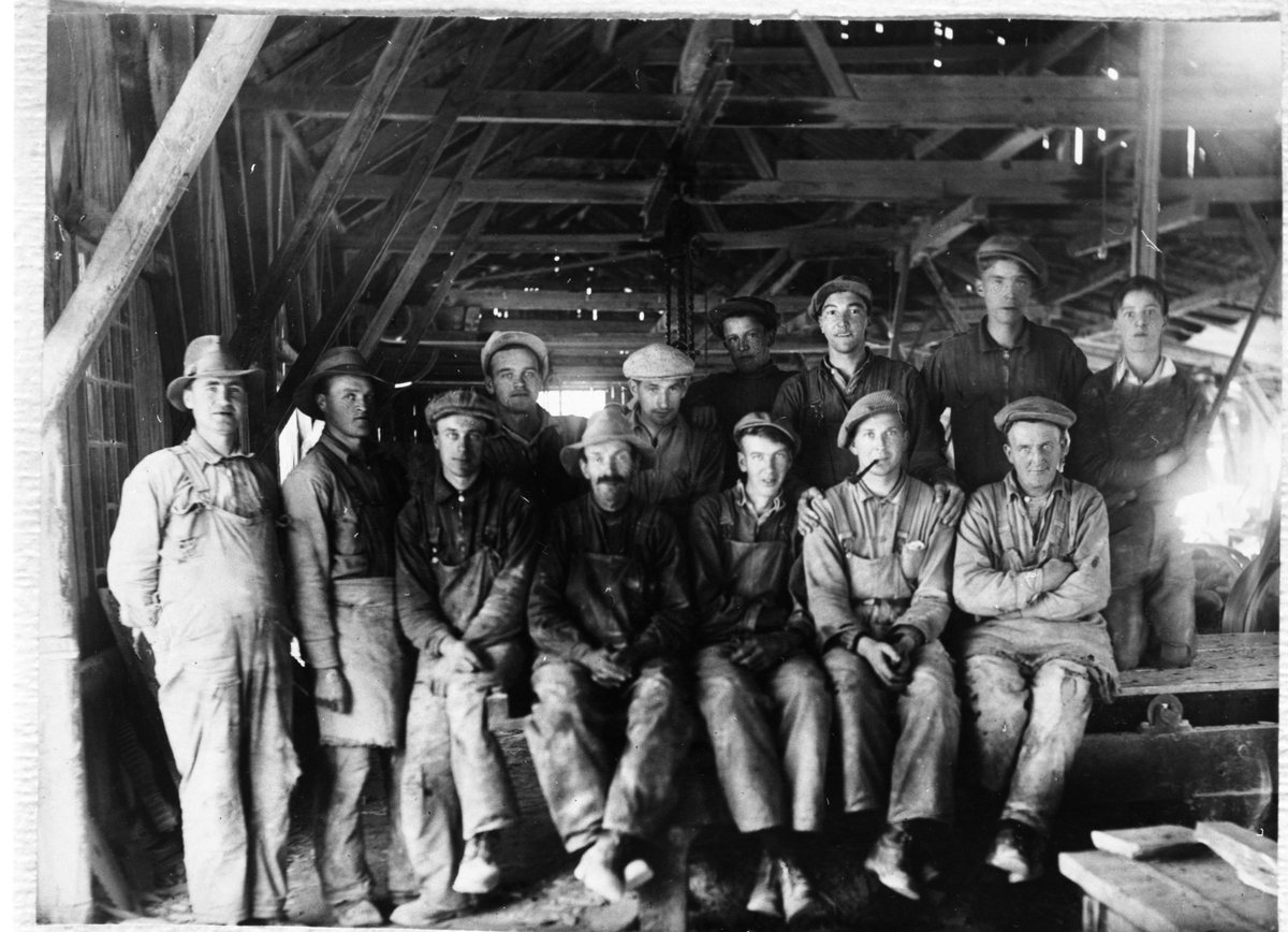 Gruppebilde av flere menn sittende inne i et av arbeidshusene ved Valdres Skiferbrud. Mennene er iført arbeidsklær og de fleste av dem også hatt og lue.