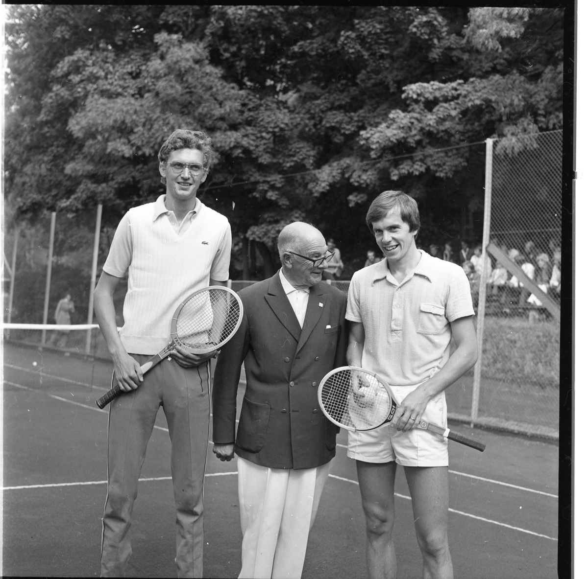 En man i ljusa byxor och mörk dubbelknäppt blazer står mellan två yngre män med tennisrack på en tennisplan.
