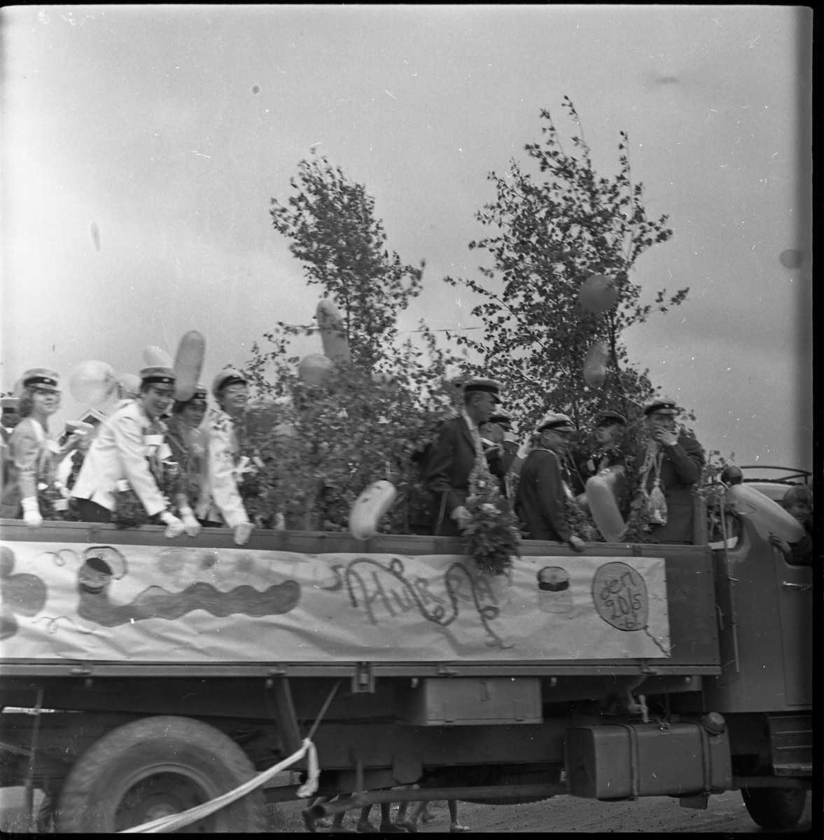 Realexamen 1961. Bilden föreställer ett lastbilsflak, på flaket står flera studenter. De bär studentmössa och blombuketter runt sina halsar. Flaket är klätt med Björkris och ballonger.